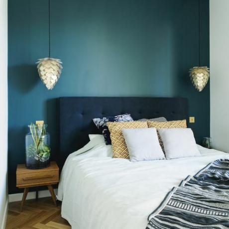 Elegant sovruminredning med litet trä nattbord, trädgård i en burk, vita sängkläder, färger pilows och filt. Utrymme med blå väggar och brun träparkett. Designlampa.