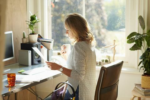 Kvinna som dricker kaffe och läser pappersarbete på skrivbordet i soliga hemmakontor.