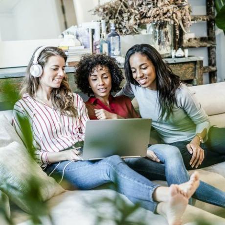 Tre lyckliga kvinnor med bärbar datorsammanträde på soffan