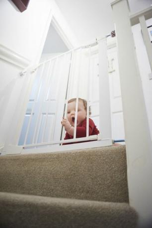 Babysäkerhetsgrind på trappan: pojke vid landningen av sitt hem. Han är bakom en babysäkerhetsport som hindrar honom från trappsteget.